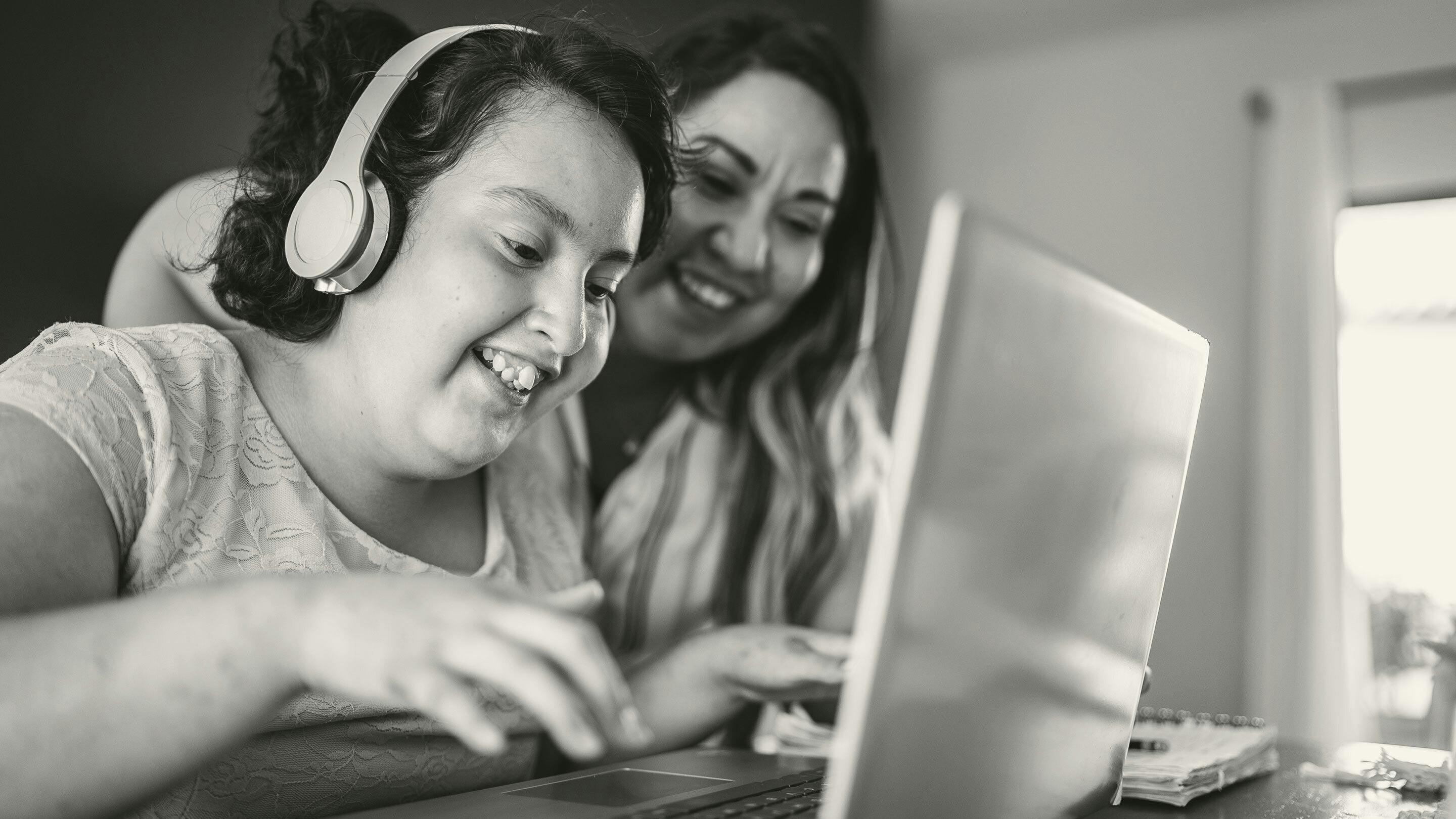 Disabled high schooler online with headphones on smiling and doing schoolwork on a laptop, with mother in background helping
