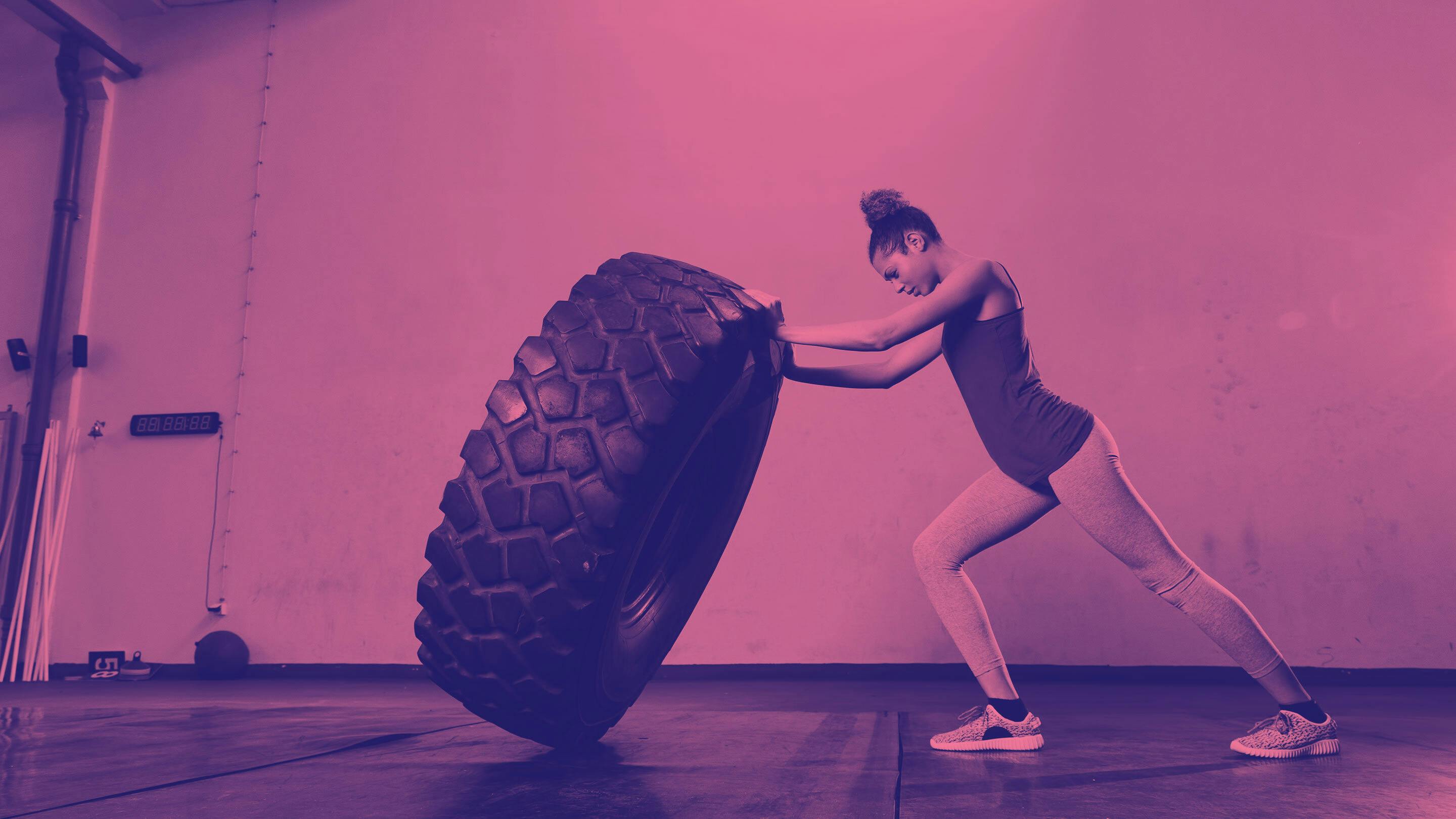Woman in athletic gear pushing large tire over in gym